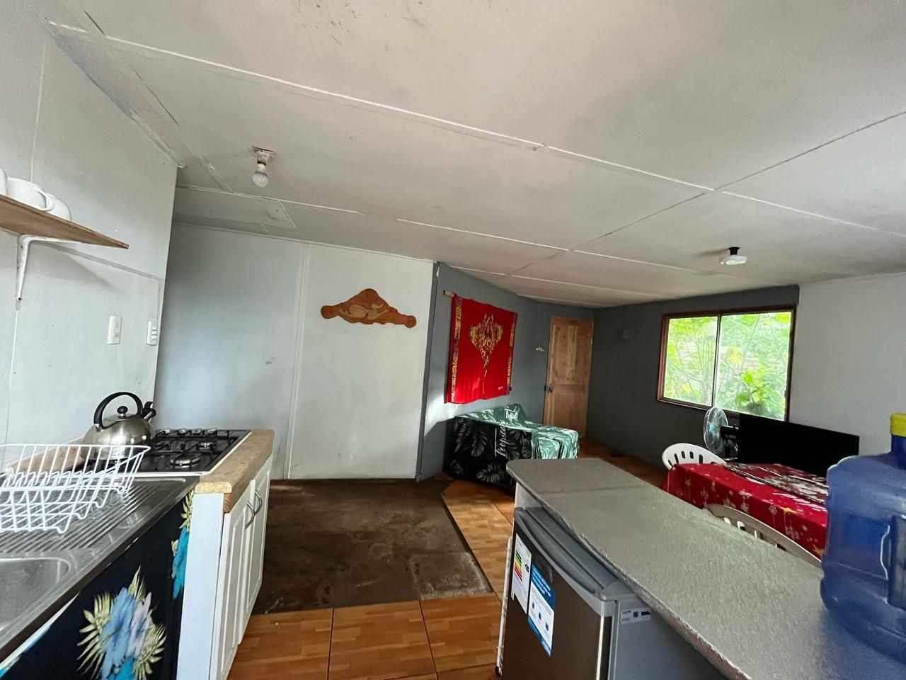 A compact kitchen with a gas stove, kettle, and a small refrigerator. A dining area with a table and chairs is visible, and a window provides natural light.