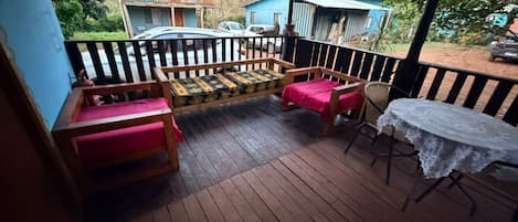 Un área de estar al aire libre con techo, muebles de madera y una mesa, con vistas a una calle residencial.