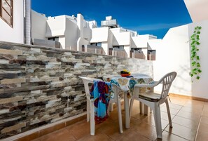 Eine Terrasse mit einer Steinmauer, einem Tisch mit Stühlen und Blick auf weiße Gebäude unter blauem Himmel.