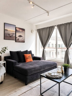 Un salon moderne avec un canapé gris foncé, deux tableaux encadrés au mur, une fenêtre avec des rideaux gris et une petite table sur laquelle est posée une plante.