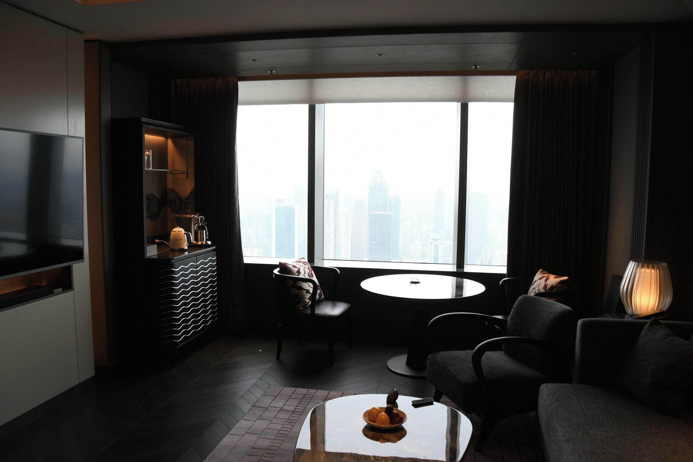 A modern hotel room with a large window, a flat-screen TV, a sofa, a dining table, and a minibar.