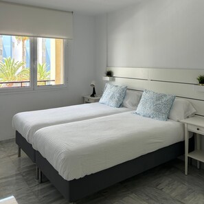 A modern bedroom with a large bed, a bedside table with a lamp, and a window with a view of palm trees. - 2 Bedroom apartment in Los Cristianos K210 (Oasis del Sur)