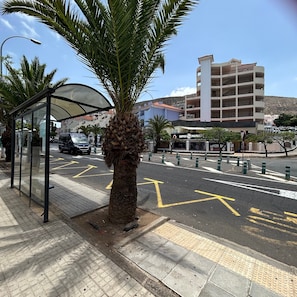 A bus stop with a palm tree, a building with multiple floors, and a parking area with directional arrows. - 2 Bedroom apartment in Los Cristianos K210 (Oasis del Sur)