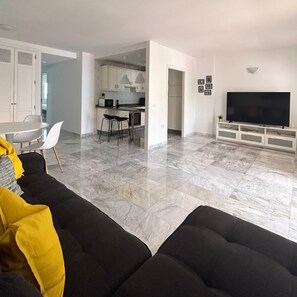 Uma sala de estar moderna com um sofá preto, uma área de jantar com cozinha ao fundo e uma TV sobre um suporte branco.