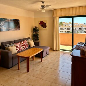 A living room with a sofa, a coffee table, a ceiling fan, a large window with curtains, and a view of buildings outside. - Vacation Home 'Apt with Ocean View San Blas K104' with Wi-Fi (Golf del Sur)