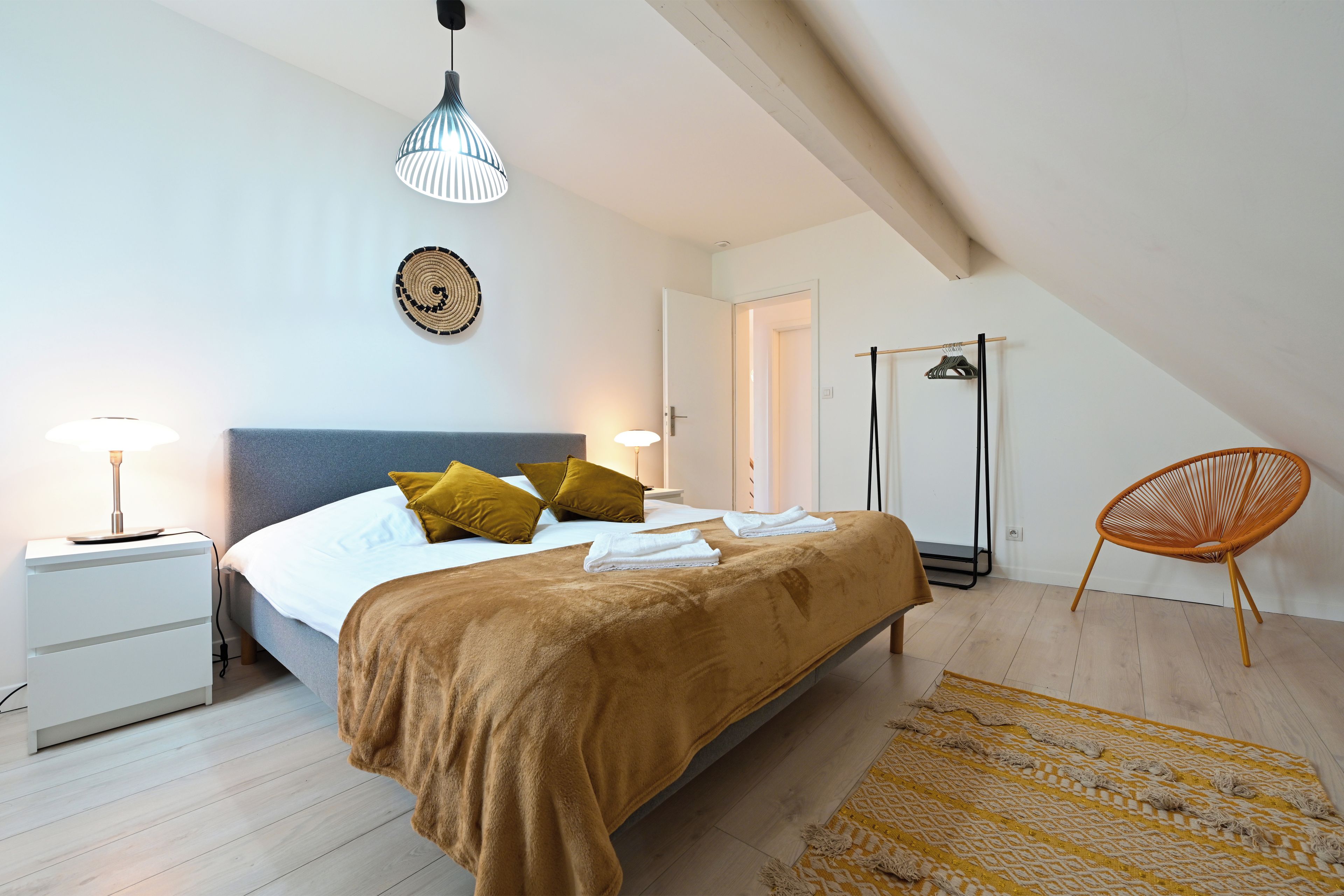 A bedroom with a bed, bedside tables, a hanging light fixture, a wall hanging, a door, a chair, and a patterned rug.