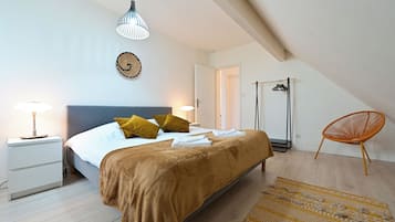 A bedroom with a bed, bedside tables, a hanging light fixture, a wall hanging, a door, a chair, and a patterned rug.