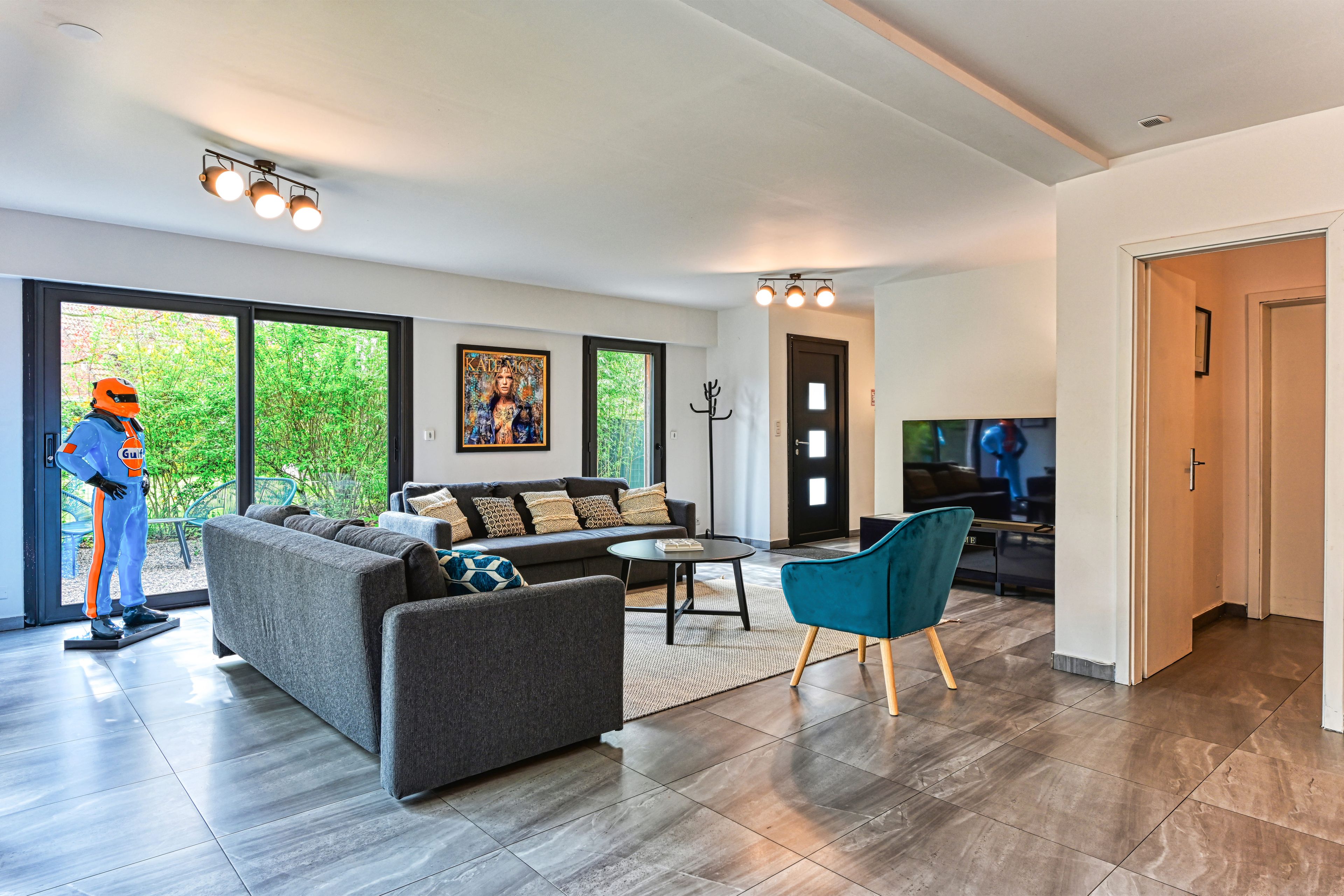A modern living room with a grey sofa, a teal armchair, and a coffee table. There is a large window with a view of greenery outside.