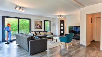 A modern living room with a grey sofa, a teal armchair, and a coffee table. There is a large window with a view of greenery outside.