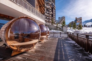 Eine Holzterrasse mit klaren, zylindrischen Strukturen, einem Holenzaun und einer verschneiten Landschaft mit Gebäuden im Hintergrund.