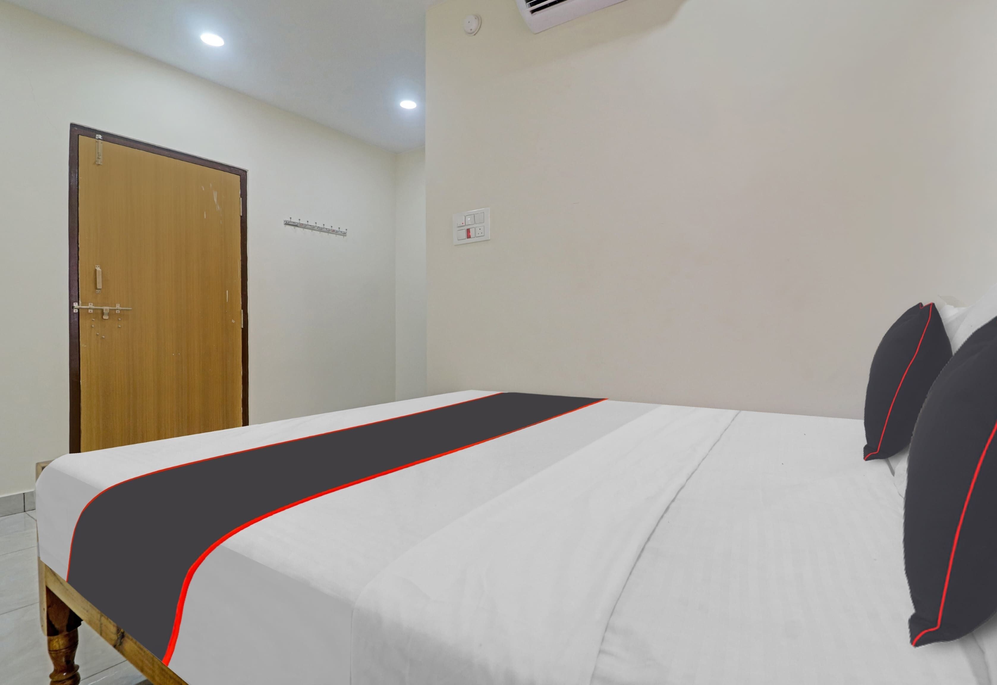 Habitación estándar de hotel con una cama individual, una puerta de madera y una pared blanca con interruptor de luz y detector de humo.