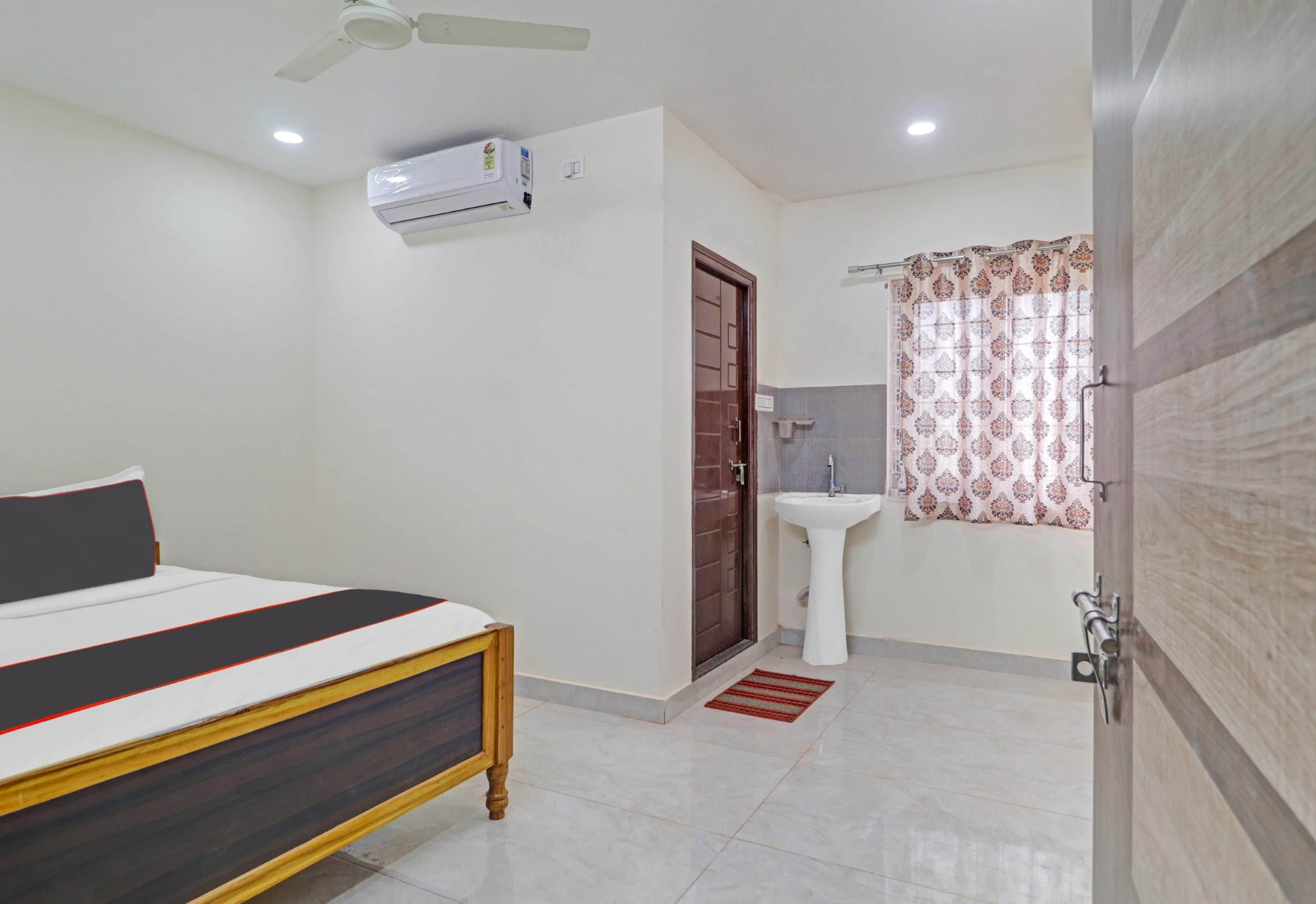Un dormitorio con cama, ventilador de techo, equipo de aire acondicionado, puerta, baño con lavamanos, espejo y cortina estampada.