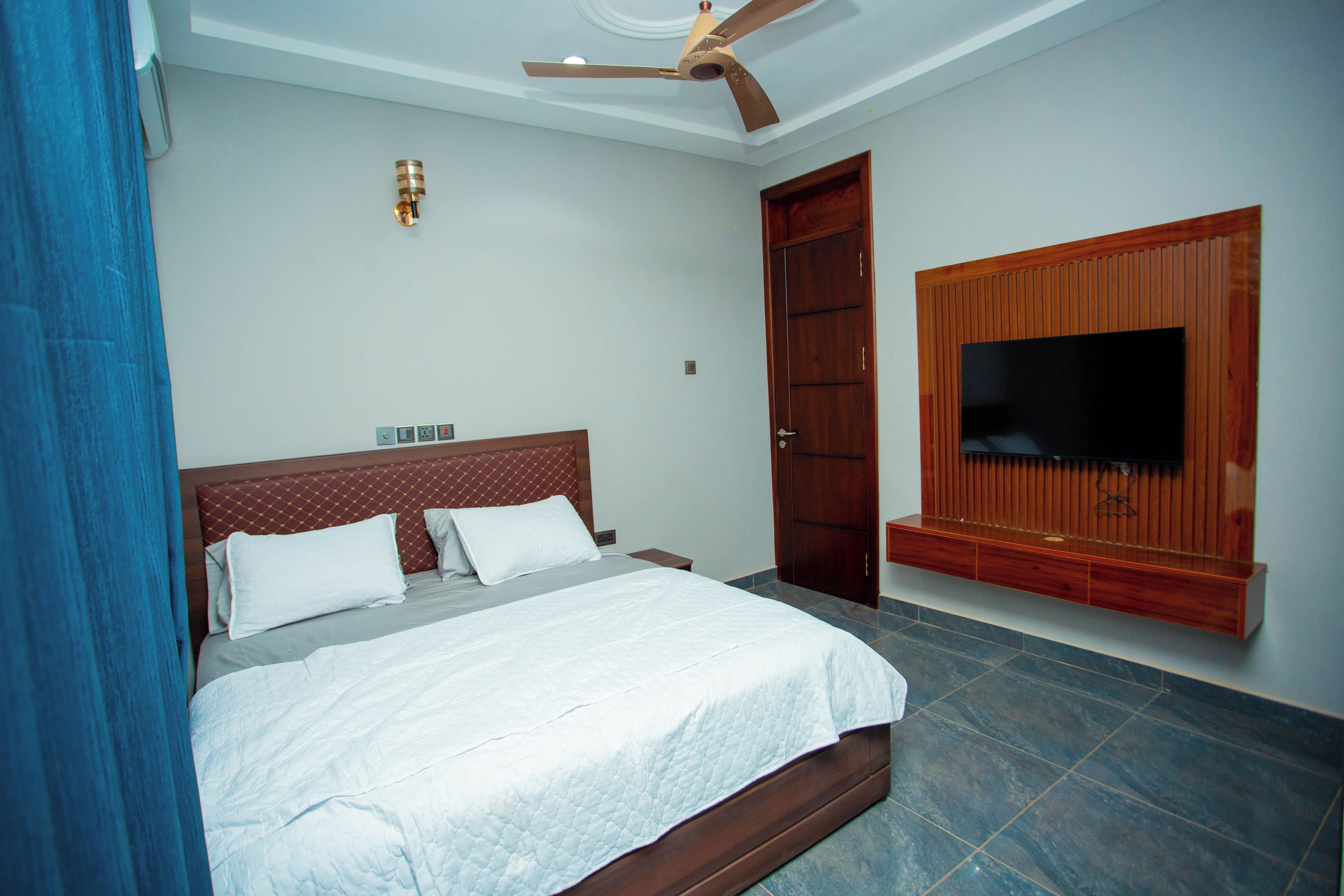Un dormitorio con cama, ventilador de techo, televisor montado en la pared y puerta de madera.
