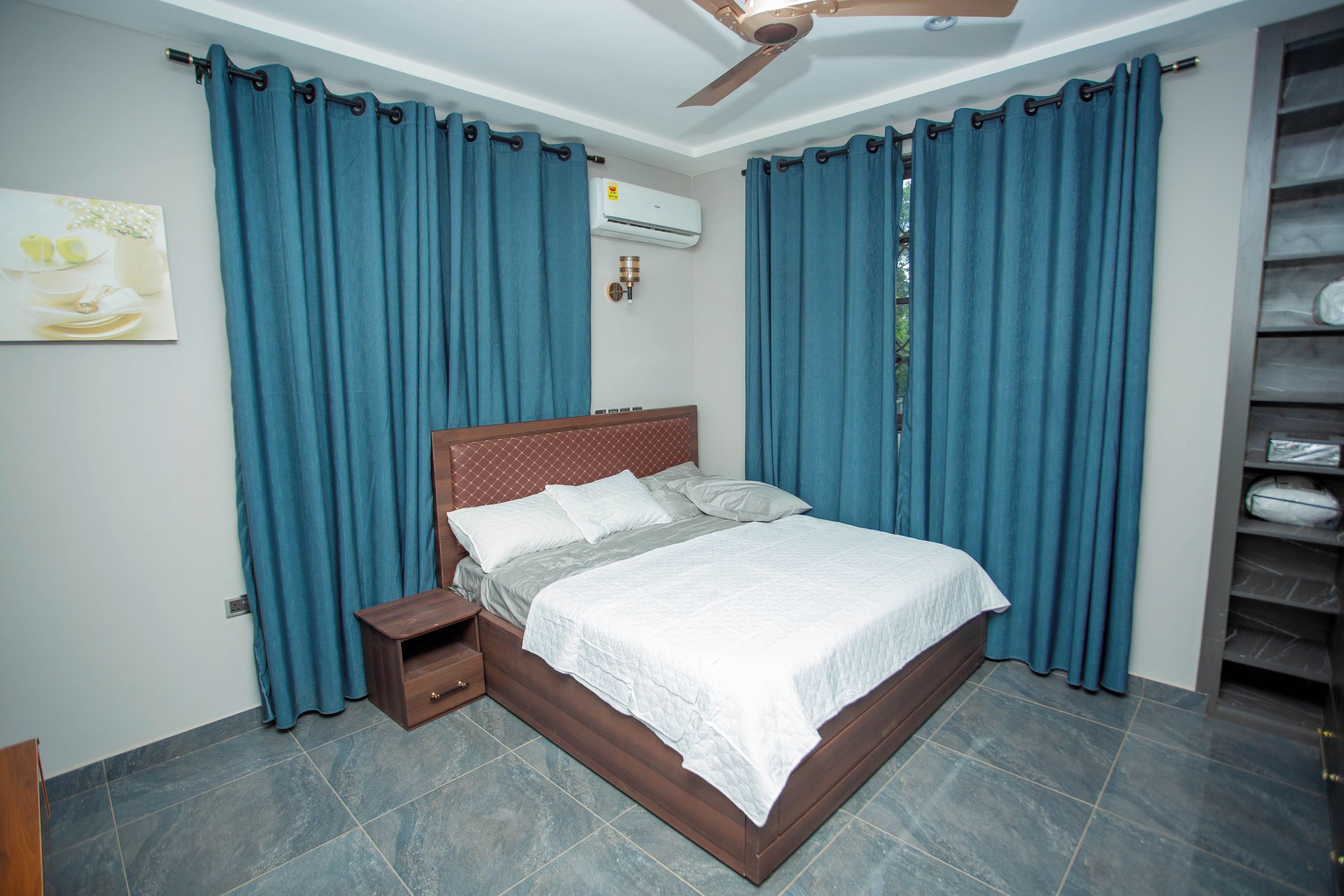 A bedroom with a bed, curtains, an air conditioning unit, a nightstand, and a wall-mounted shelf.