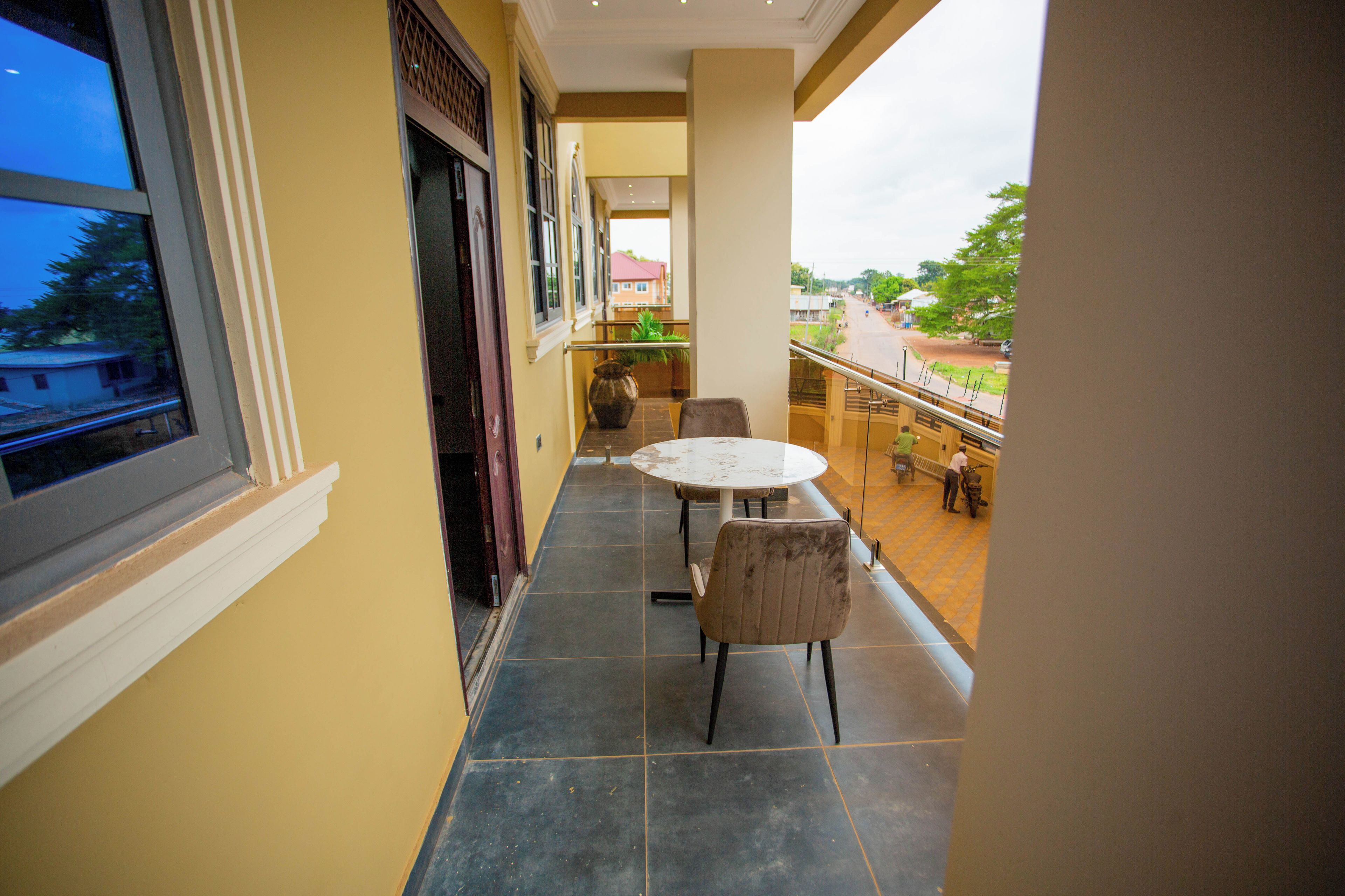 A narrow balcony with a table and chairs, a view of the street, and a large vase on the floor.
