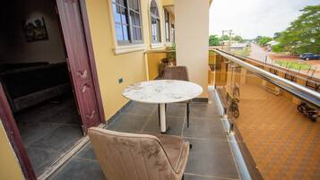 A balcony with a round table and two chairs, overlooking a street with parked cars and greenery.