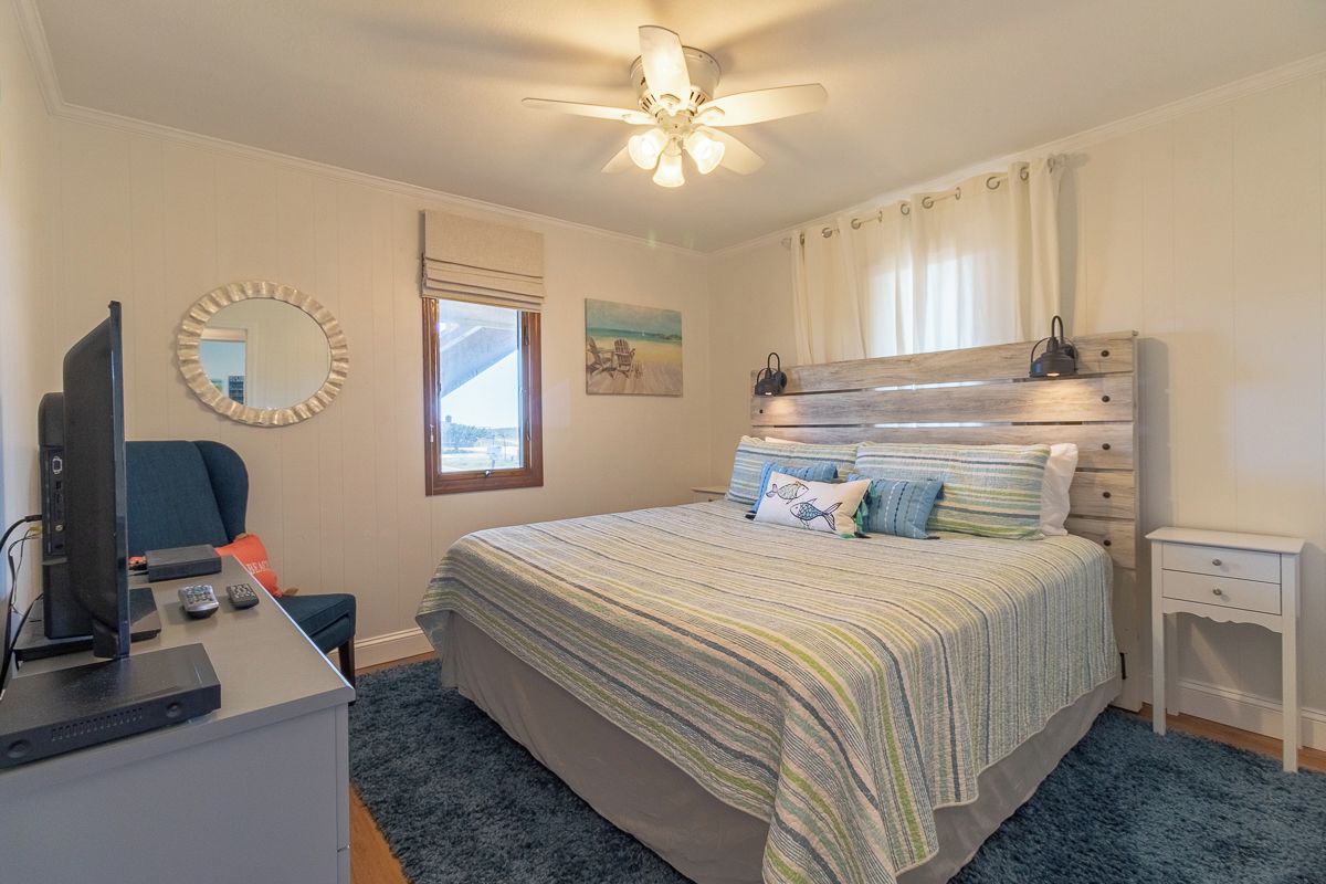 Un dormitorio con cama, escritorio con televisor, ventilador de techo y ventana con cortinas.