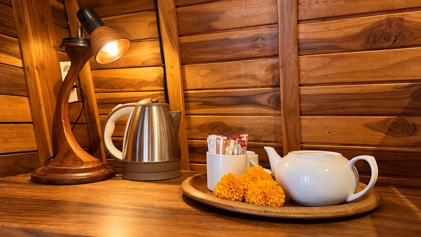 A wooden table with a lamp, a kettle, a mug, and a tray with flowers.
