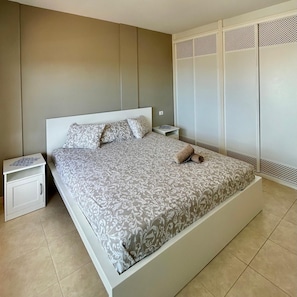 A bedroom with a large bed, white nightstands, a small table, and a wardrobe with a louvered design. - Vacation Home 'Sea View 1 Bedroom Chayofa K109' with Mountain View, Shared Pool and Wi-Fi (Arona)