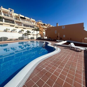 A swimming pool with a tiled deck, surrounded by a low wall and palm trees. - Vacation Home 'Sea View 1 Bedroom Chayofa K109' with Mountain View, Shared Pool and Wi-Fi (Arona)