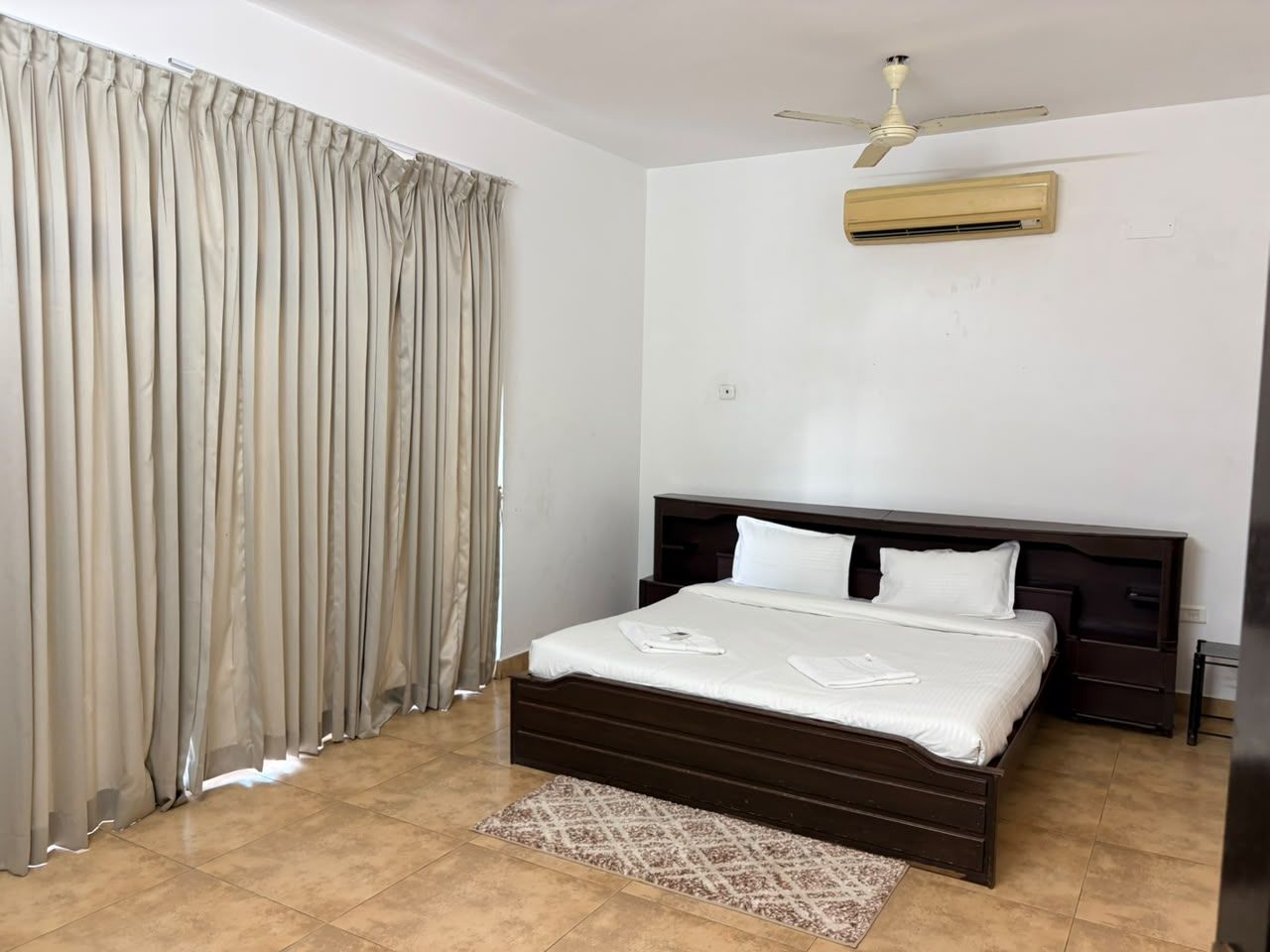 A bedroom with a bed, curtains, a ceiling fan, an air conditioning unit, and a rug.