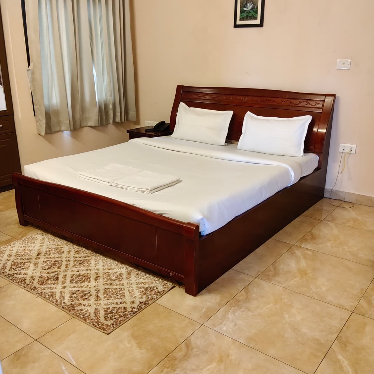 Cama bem arrumada com cabeceira de madeira, tapete estampado e parede lisa com um único quadro.