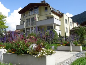 En bygning med flere etager og altaner, omgivet af blomstrende planter og buske.