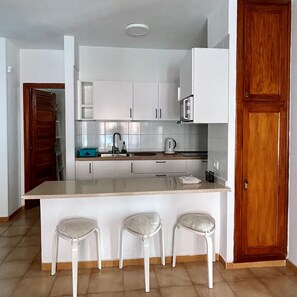 A modern kitchen with white cabinets, a central island with stools, a wooden door, and a tiled backsplash.