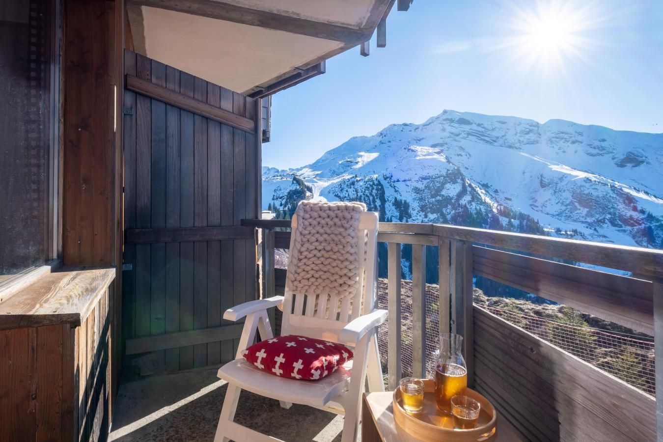 Una terraza de madera con una silla blanca y una mesa con una bebida. Se ve una vista de la montaña al fondo.