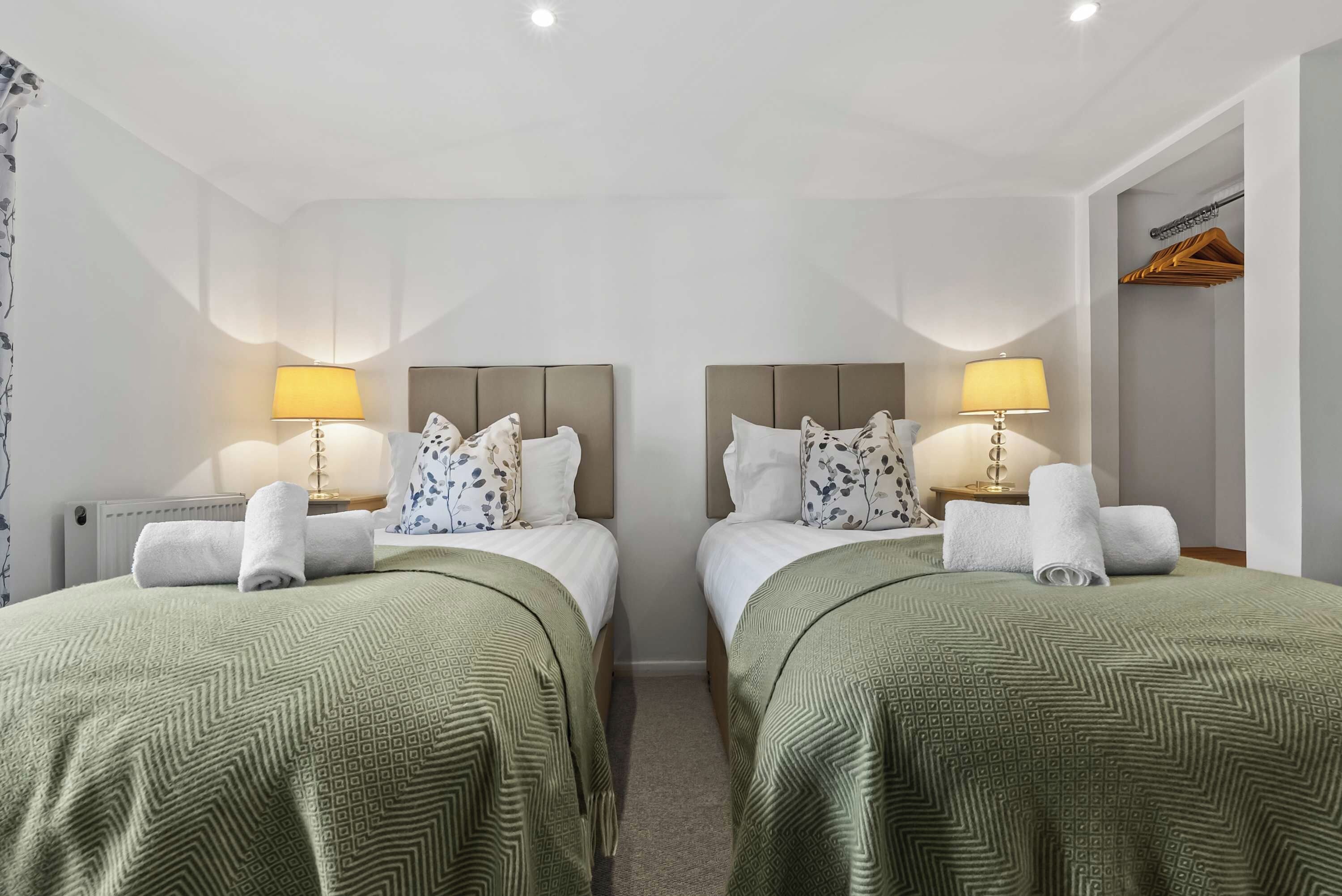 Habitación de hotel con dos camas, cada una con una colcha verde estampada, almohadas blancas y un cabecero. Hay dos lámparas de noche con sus respectivas pantallas, un radiador y un perchero de madera en la pared.
