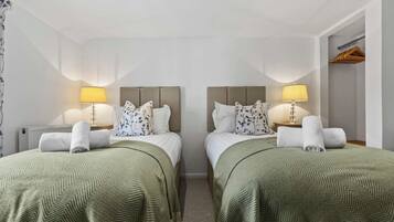 Habitación de hotel con dos camas, cada una con una colcha verde estampada, almohadas blancas y un cabecero. Hay dos lámparas de noche con sus respectivas pantallas, un radiador y un perchero de madera en la pared.