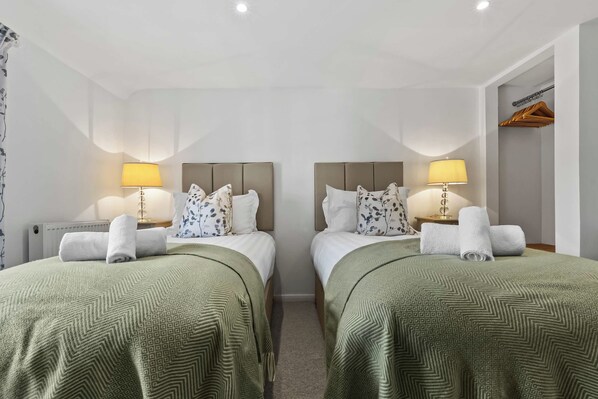 Habitación de hotel con dos camas, cada una con una colcha verde estampada, almohadas blancas y un cabecero. Hay dos lámparas de noche con sus respectivas pantallas, un radiador y un perchero de madera en la pared.