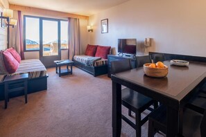 Ett hotellrum med en säng, soffa, TV, matbord och utsikt över bergen genom fönstret.