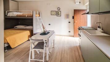 A compact room with a bunk bed, a small dining table with a chair, a kitchen area with a sink and stove, and a wooden door.