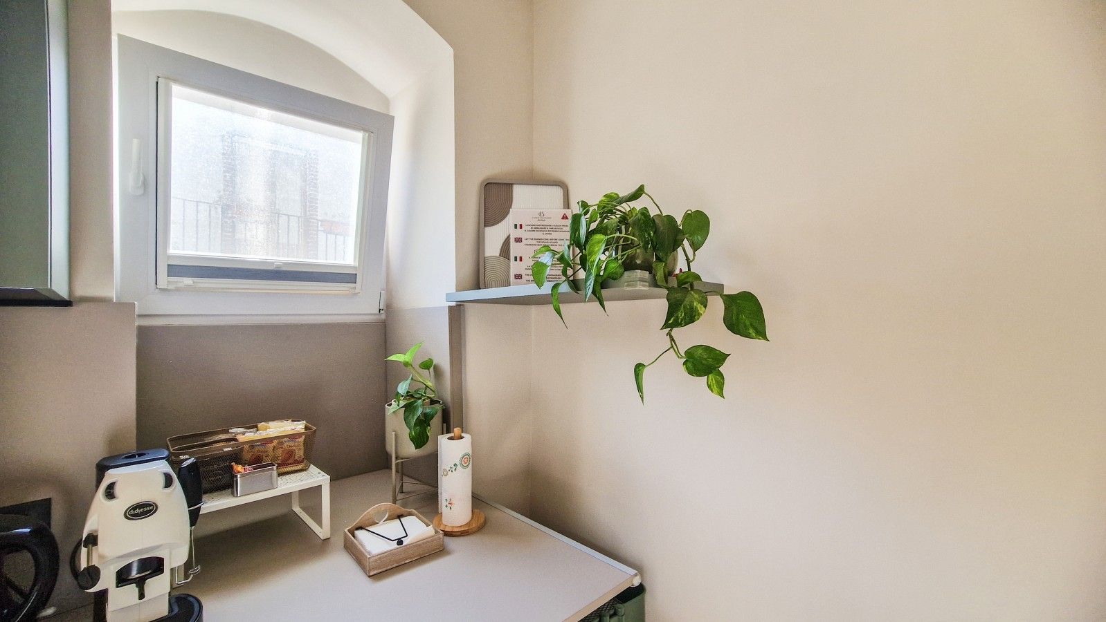 Une pièce d’angle avec une fenêtre, une petite étagère remplie de livres, une plante et un bureau avec une bouilloire et une boîte.