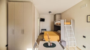 Habitación de hotel compacta con una cama litera, un escritorio con silla, un armario, un televisor y una mesita de noche con un florero.