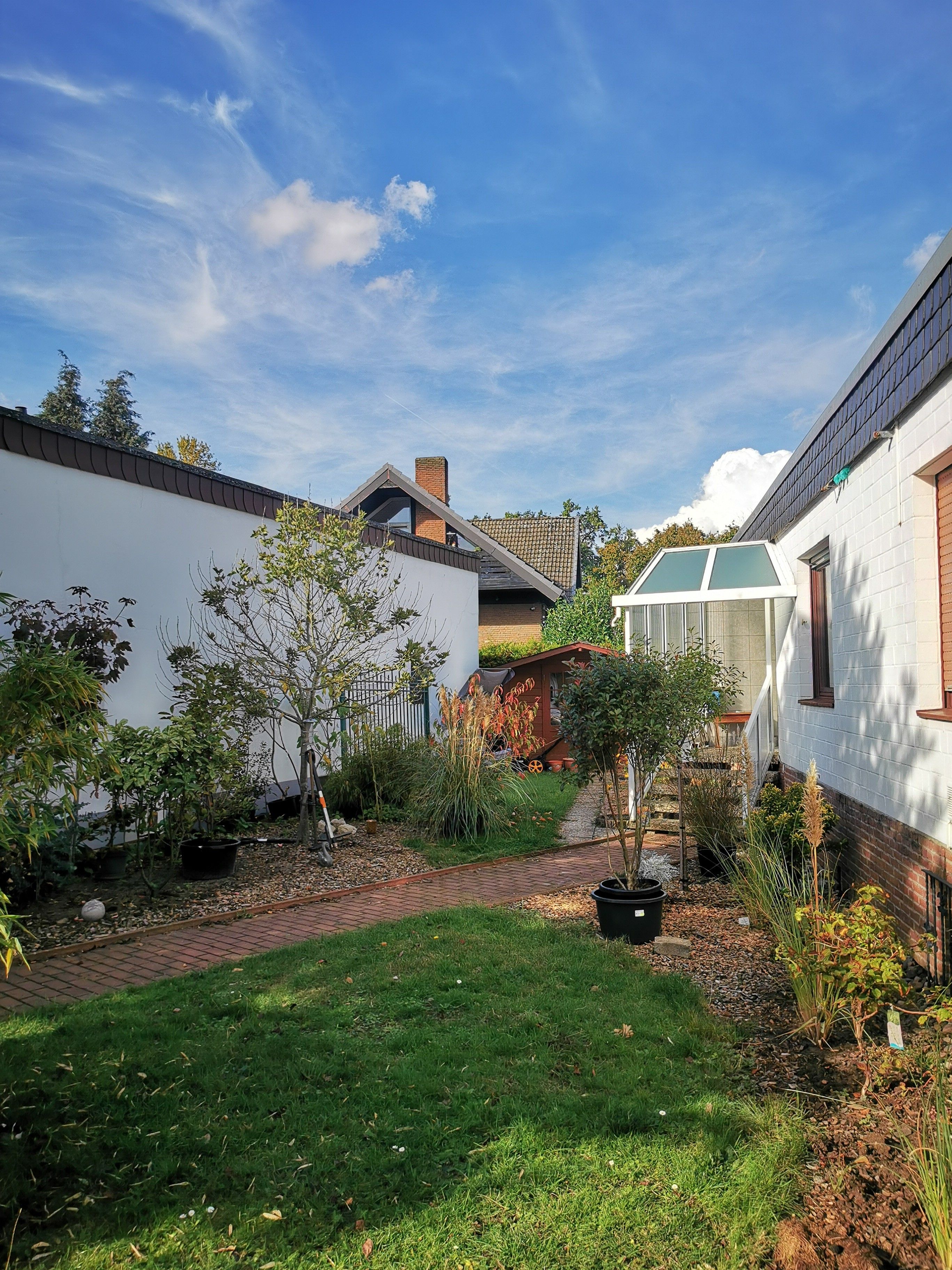 A residential garden with a paved pathway, various plants, and a white building with a green roof.