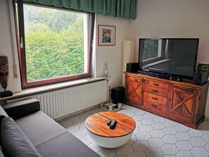 En stue med et stort vindue, et fladskærms-tv, et træskab og et rundt sofabord.