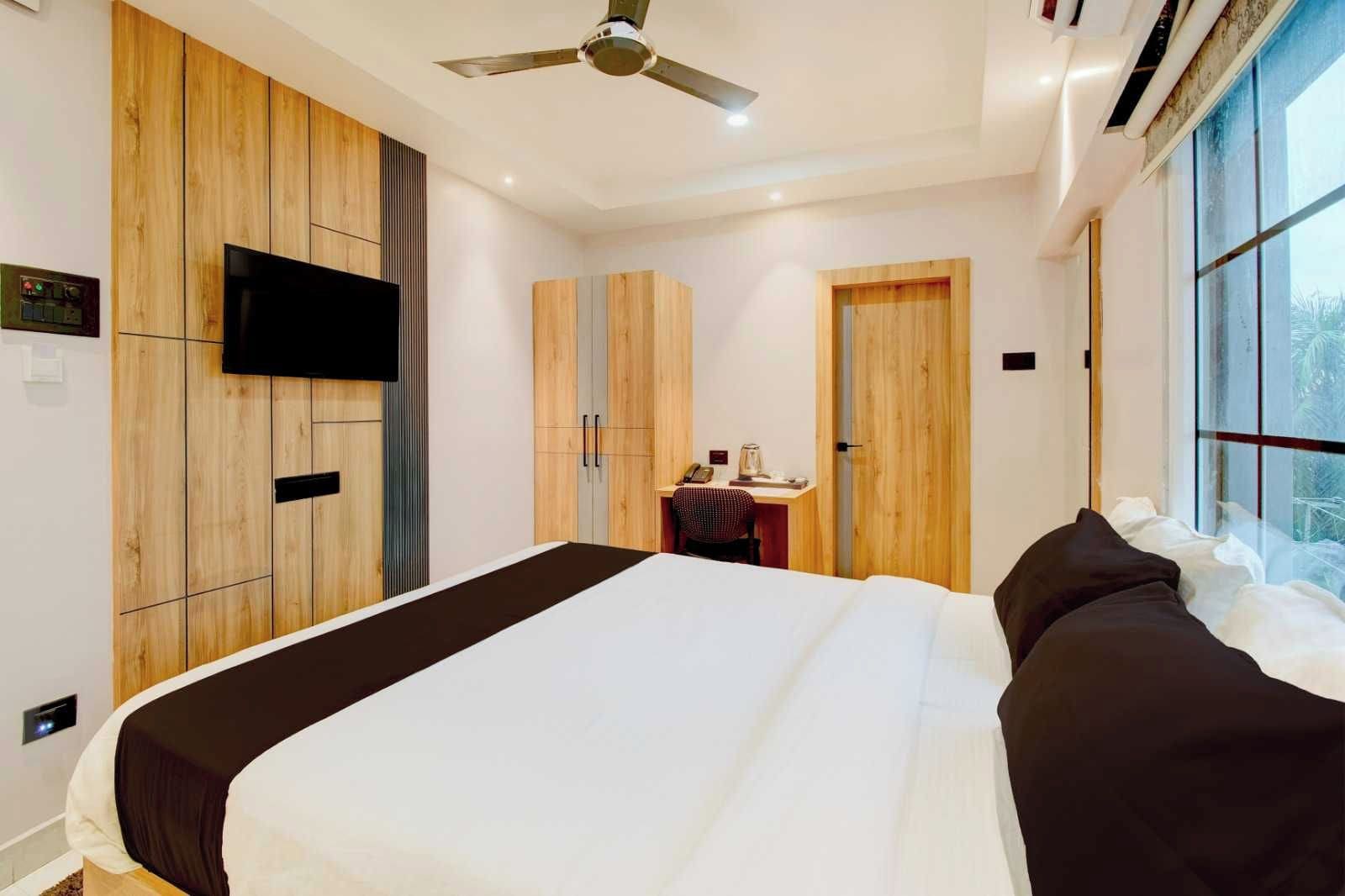 A hotel room with a large bed, wooden wardrobe, a desk with a chair, a television mounted on the wall, and a ceiling fan.
