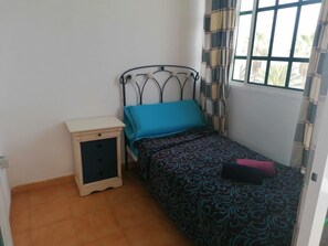 Ein kleines Zimmer mit einem Einzelbett, einem Metallkopfteil, einem Nachttisch mit Schubladen und einem fenster mit Vorhängen.
