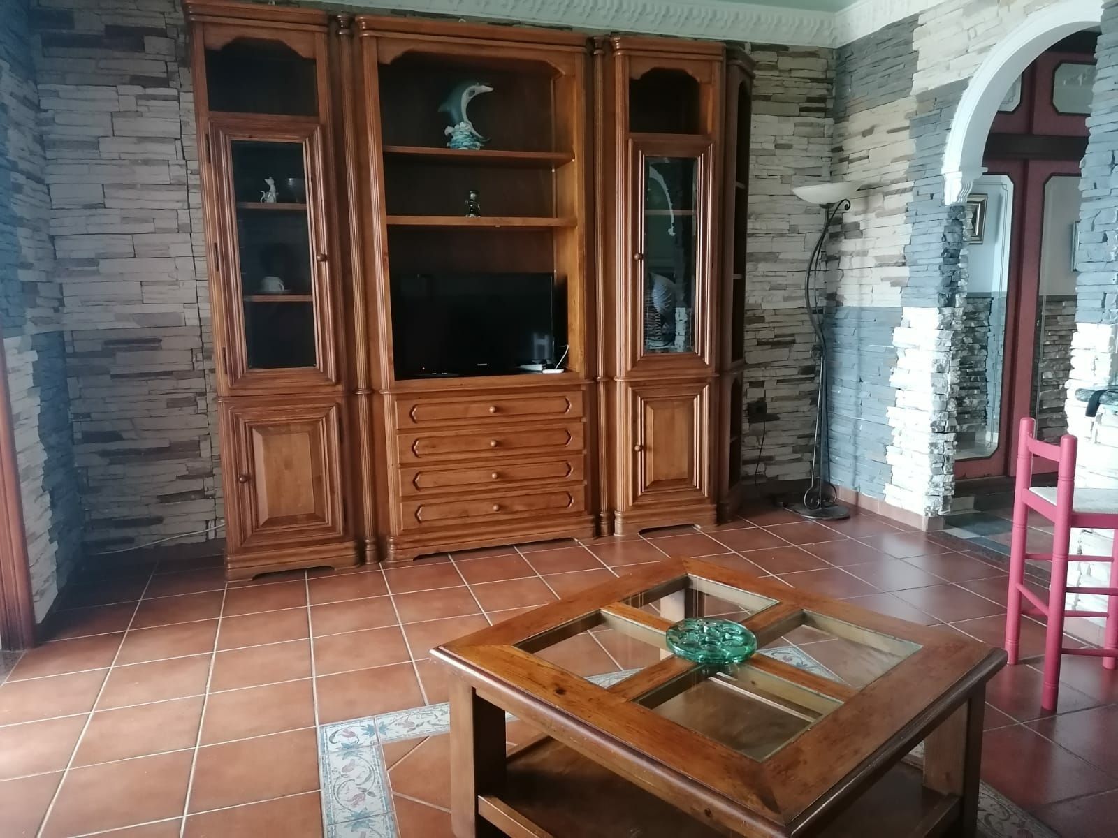Una habitación con un gran armario de madera con cajones y estantes, una mesa de centro con tapa de vidrio y un objeto decorativo, y una silla rosa.