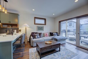 A modern living room with a sofa, coffee table, and a dining area with a kitchen counter. - Cozy Mountain Retreat- private hot tub, heated driveway, ski out (Snowshoe)