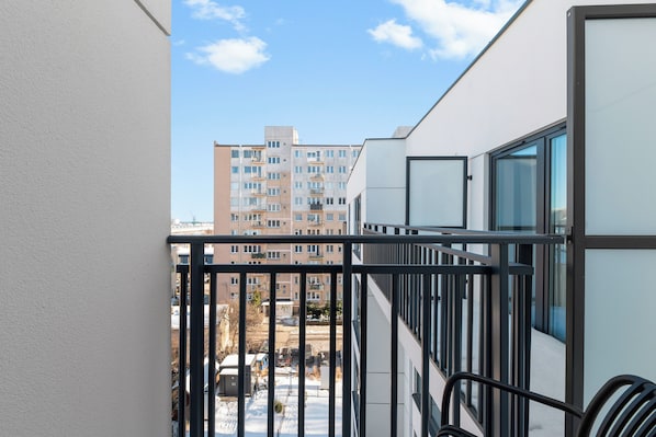 Un balcón con barandilla de metal, vista a un edificio de varios pisos y cielo despejado.