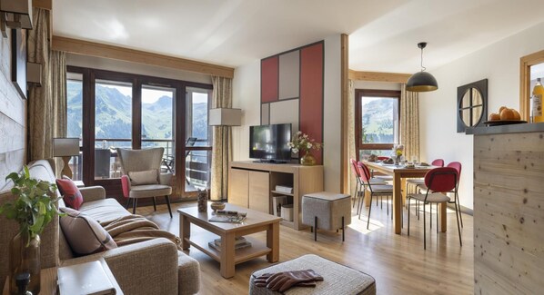 A modern living room with a sofa, coffee table, and TV. There is a dining area with chairs and a table. The room has large windows with a mountain view. - Wifi, 5m from ski slopes, view mountain, swimming pool, sauna, hammam, balcony, ski locker, 54m² (Arâches-la-Frasse)