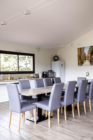 A modern dining area with a wooden table and grey upholstered chairs, a kitchen in the background, and a window with a view of greenery. - 15-person vacation rental - ALTER ET GO Eco-Lodge Village - Panoramic view of Canigou (MARQUIXANES)