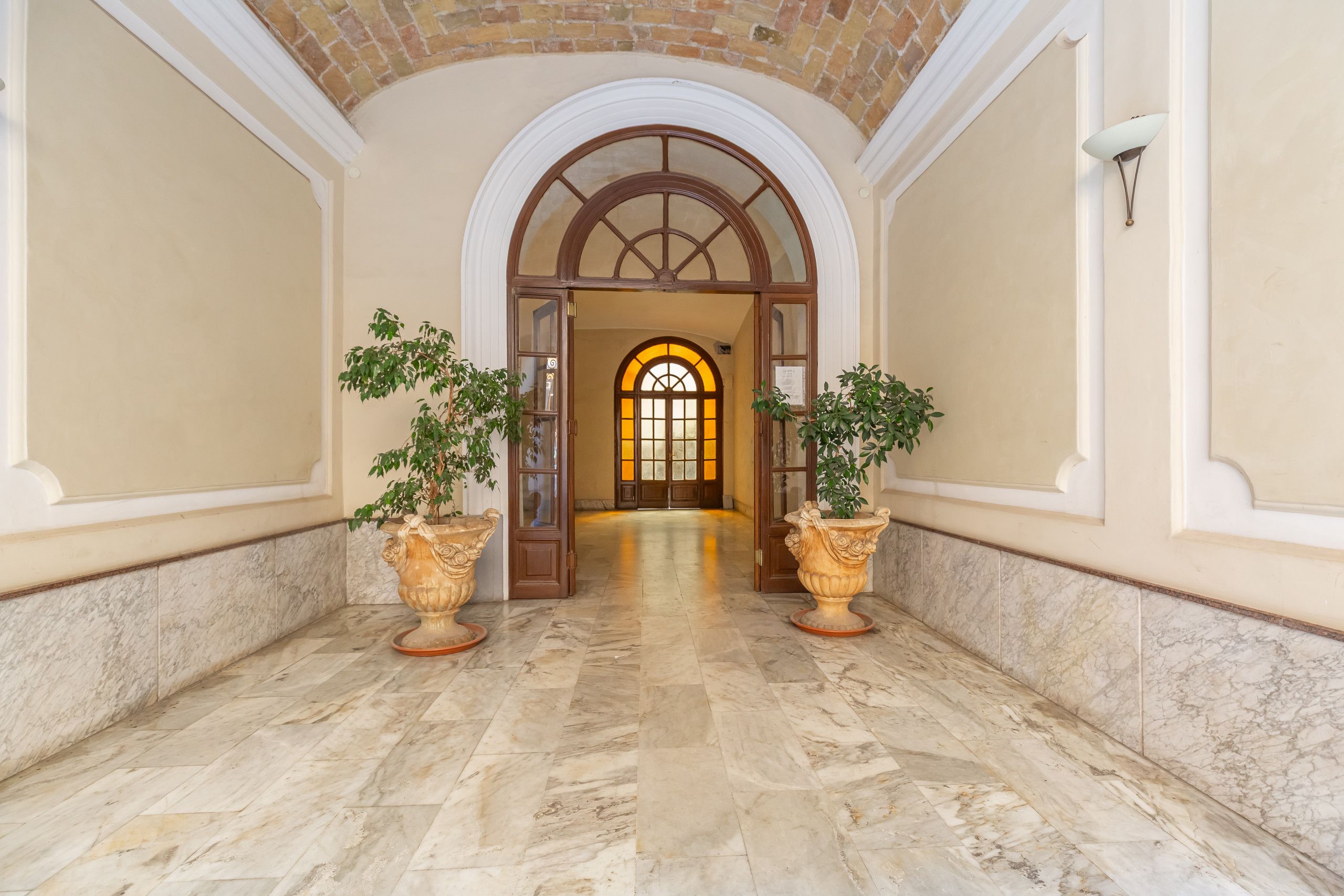 A hallway with a marble floor, two large potted plants, and an arched doorway leading to a room with a window.
