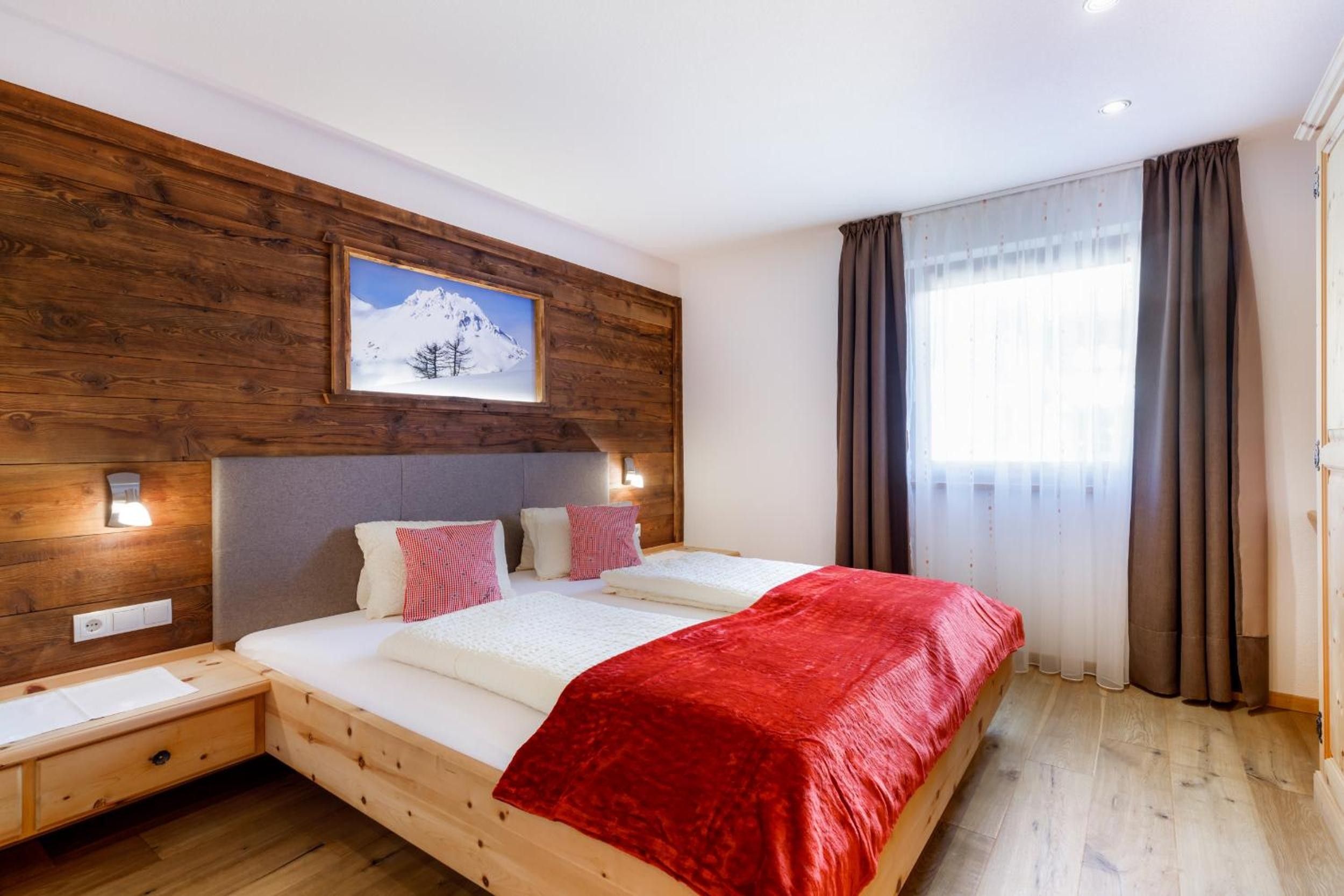 Una camera d'albergo con una testiera in legno, un letto grande con una coperta rossa, un comodino, una lampada a parete e una finestra con tende.