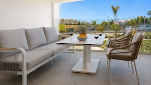 A modern outdoor seating area with a sofa, table, and chairs, overlooking a residential area with palm trees and a clear sky. - Stylish apartment with access to pool, gym & padel (Mijas Costa)
