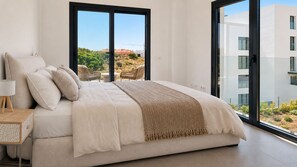 A modern bedroom with a large bed, a bedside table with a lamp, and a view of a building and landscape through large windows. - Stylish apartment with access to pool, gym & padel (Mijas Costa)