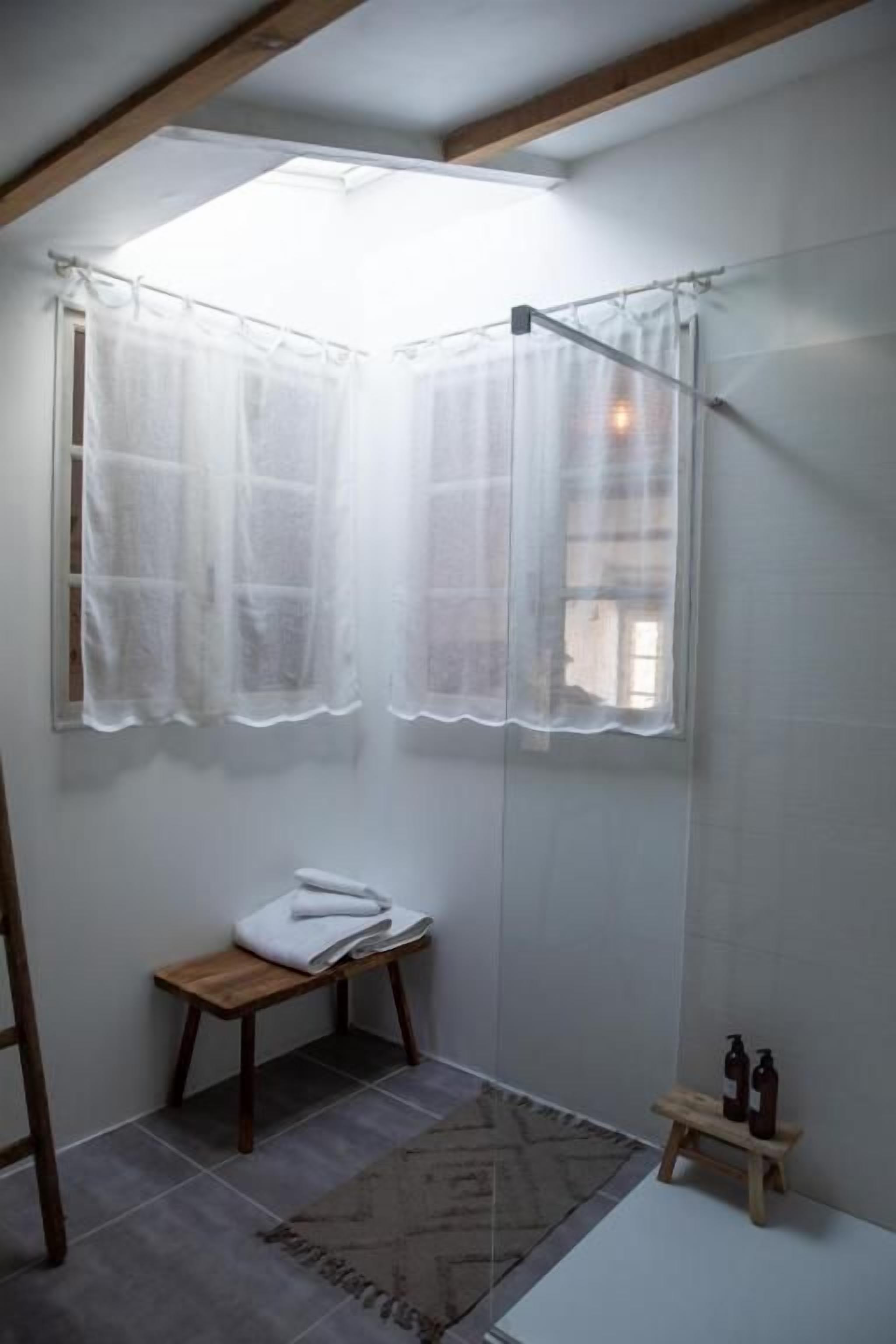 A small bathroom with a shower, a bench, and a window with sheer curtains.